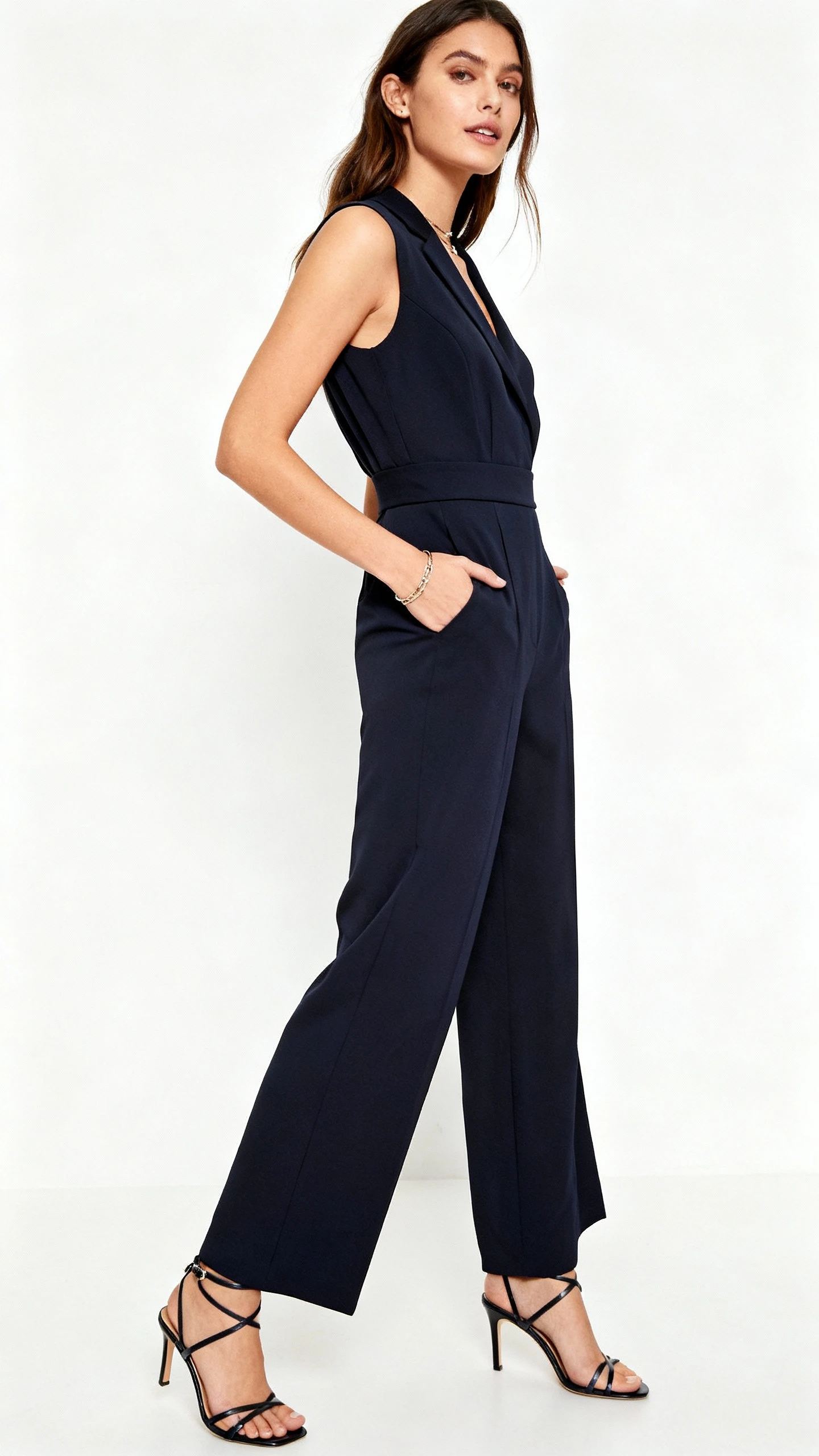 Editorial style photo of a woman wearing a luxe tailored-style jumpsuit with a defined waist and wide-leg silhouette, strappy heels, subtle jewelry, white background, studio lighting, three-quarter angle, clean and polished composition.