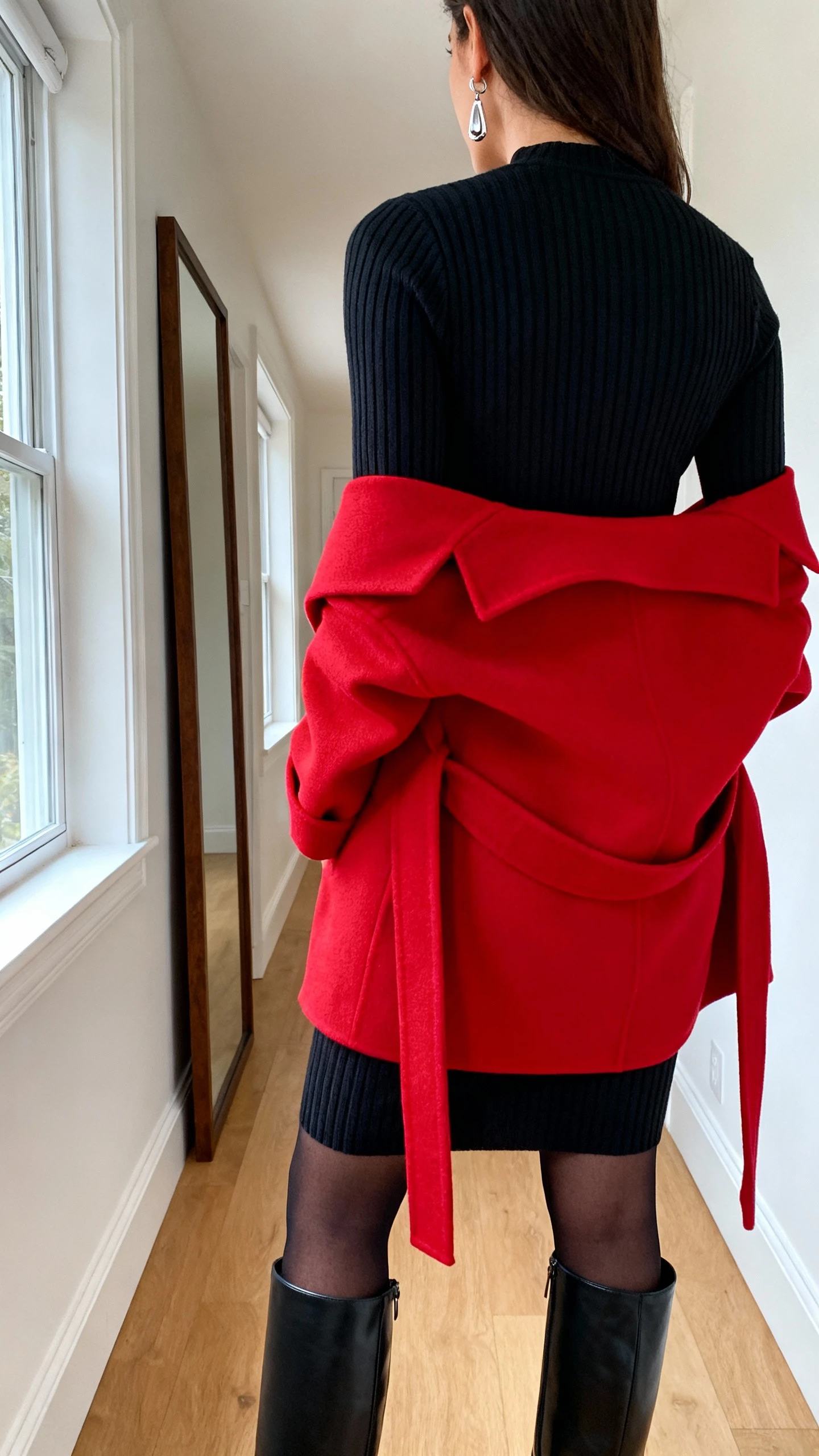 iPhone back view of a woman wearing a long-sleeve black ribbed knit dress with black opaque tights, knee-high black leather boots, a bold red wrap statement coat, and elegant drop earrings; face not visible, hallway mirror, natural daylight from window, casual iPhone photo.