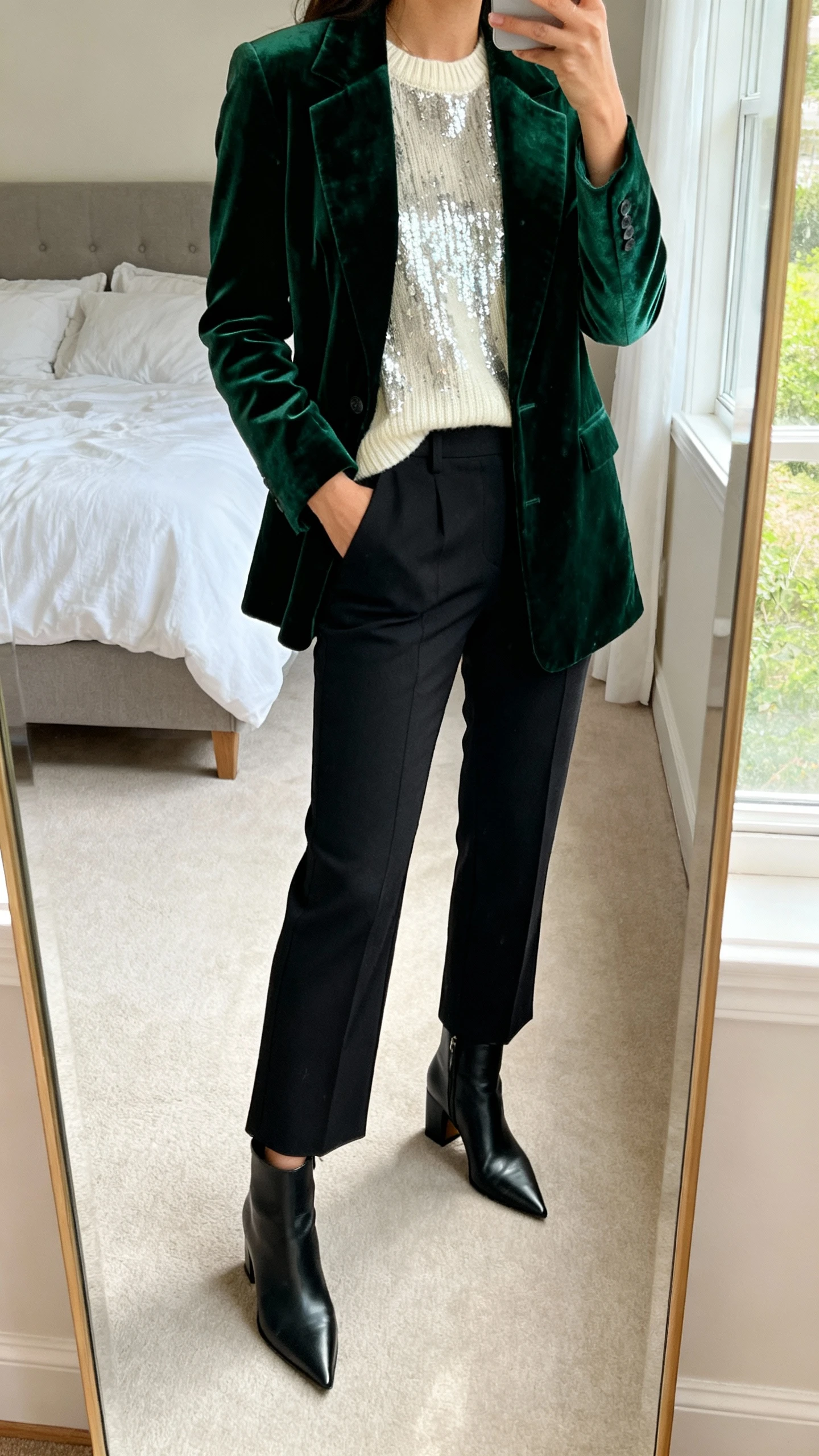 iPhone mirror selfie of a woman wearing a deep green velvet blazer over a cream metallic-thread knit top, high-waisted black cigarette trousers, and pointed-toe black block-heel ankle boots; face not visible, cropped at neck, bedroom mirror, morning window light, iPhone photo quality.