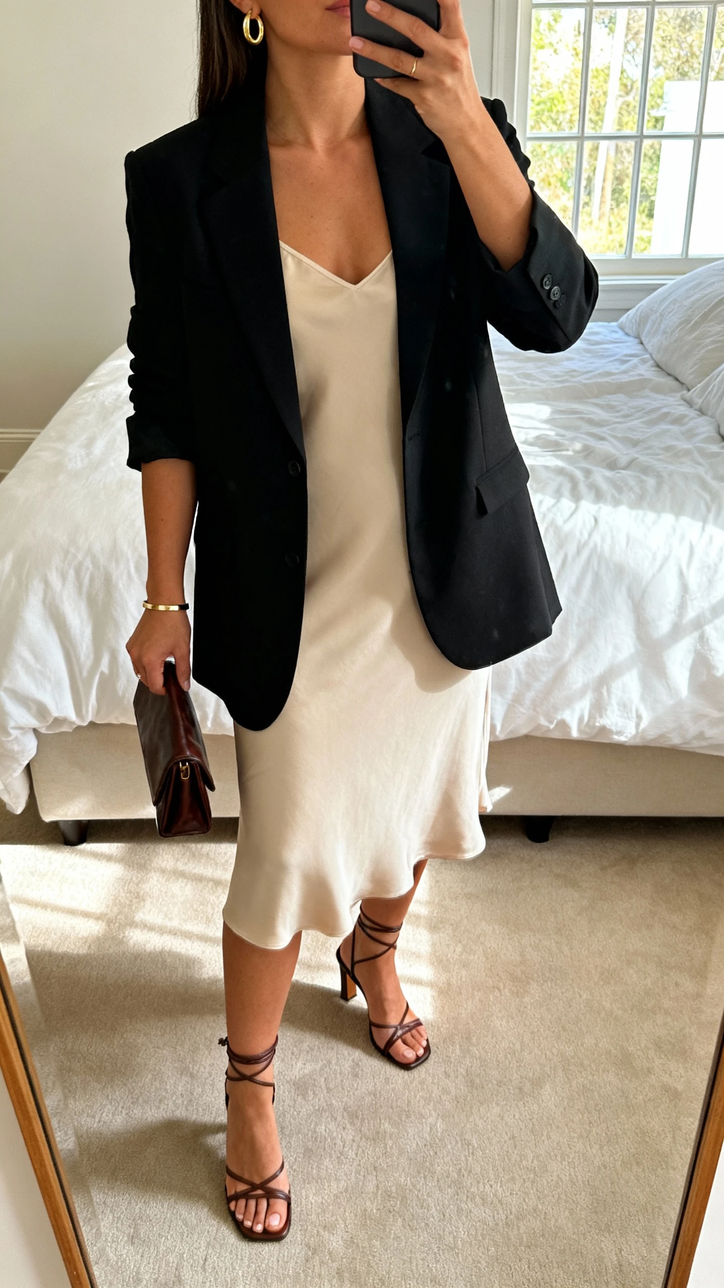 iPhone mirror selfie of a woman wearing a luxe minimal slip dress with a tailored black blazer, delicate gold hoop earrings, slim bracelet, strappy heeled sandals, and a small leather clutch, face not visible, cropped at neck, bedroom mirror with natural window light, iPhone photo quality.