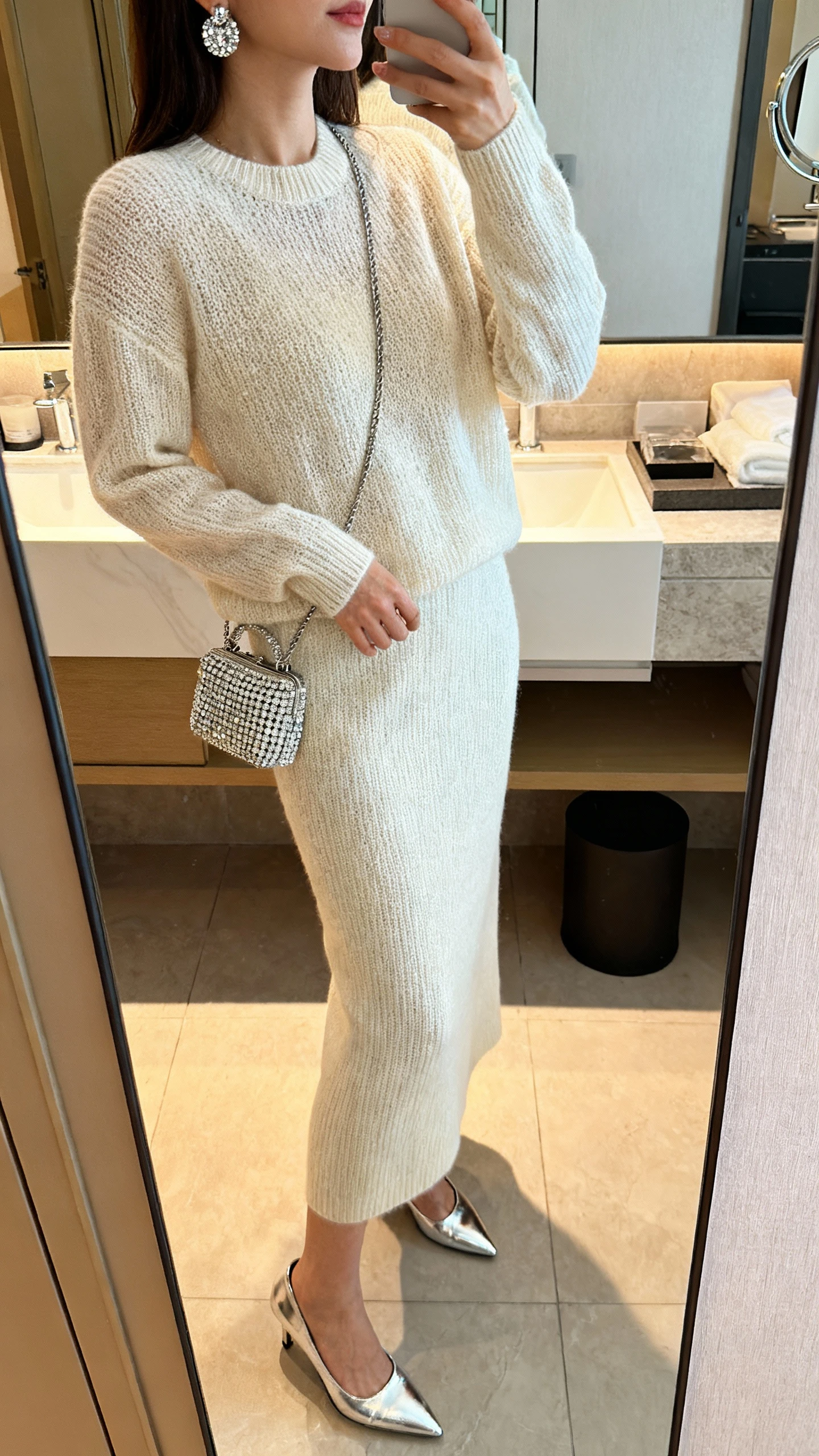 iPhone side-angle mirror selfie of a woman in a monochrome knit set (fine-knit sweater and matching knit midi skirt) with sparkly earrings, a rhinestone mini bag, and metallic heels, face not visible, bathroom full-length mirror, warm indoor lighting, iPhone photo quality.