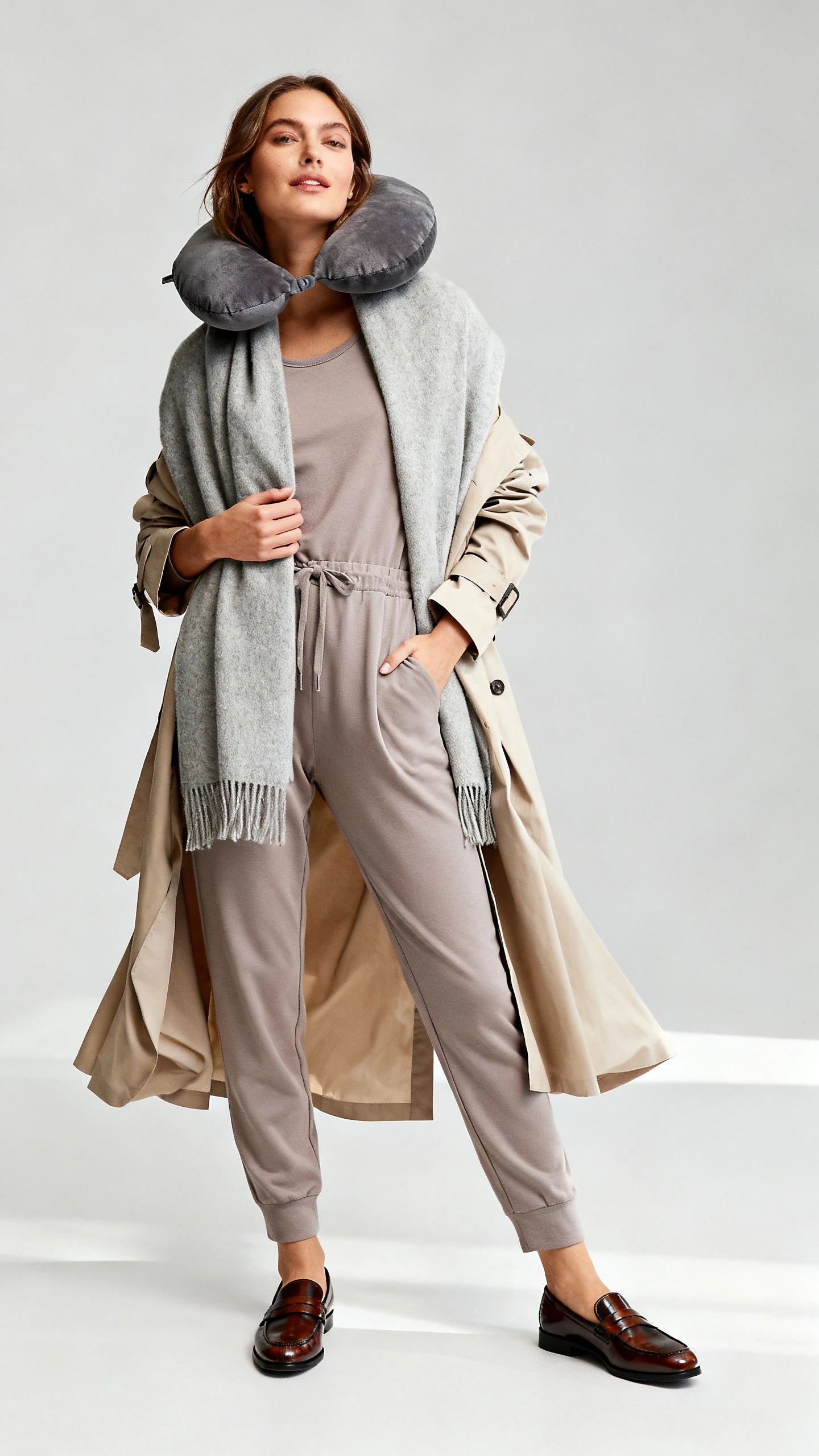 Studio fashion photo of a woman wearing a soft jersey jumpsuit layered with a cashmere wrap and lightweight trench, paired with polished slip-on loafers and oversized scarf as a travel pillow; light gray background, studio lighting, relaxed natural stance, clean composition, magazine-quality.